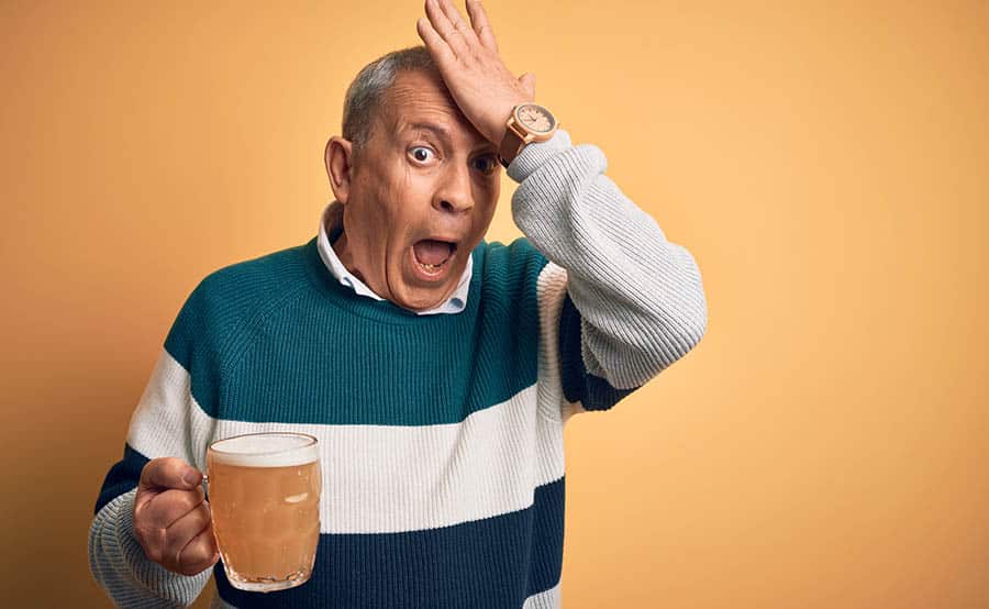 Older man with brain damage from alcohol holds a large glass of beer, with his other hand on his forehead.