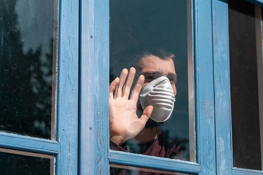 Man suffering from addiction relapse is wearing a mask and looking out the window while in covid 19 quarantine. 