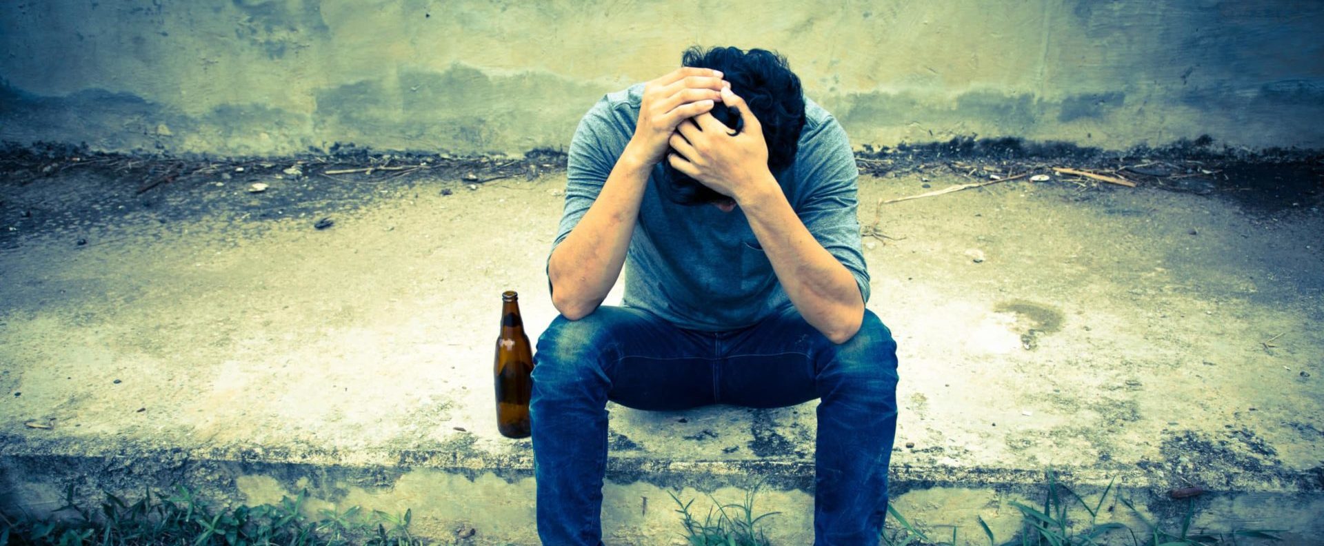 A guy is sitting on dirty cement with debris in the back, he has a beer bottle next to him and his head in his hands