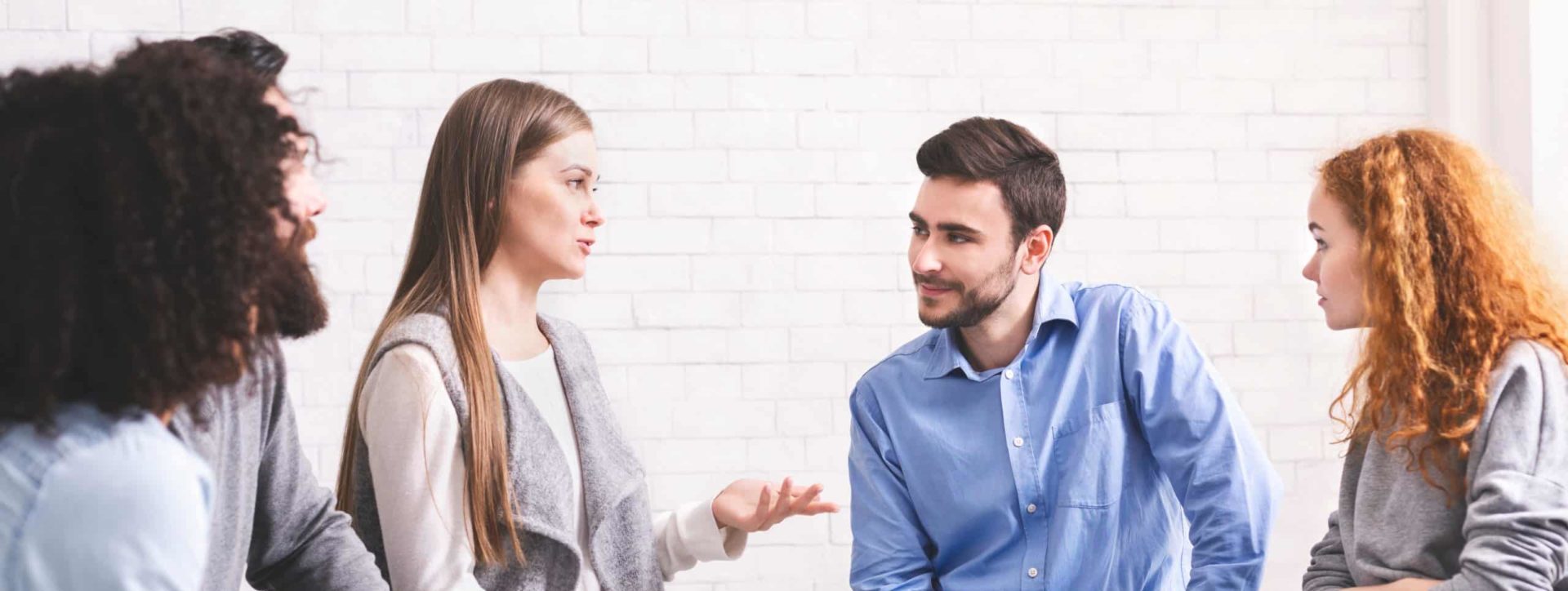 Four adults sitting in a half circle, talking to one another in brightly lit room with brick walls