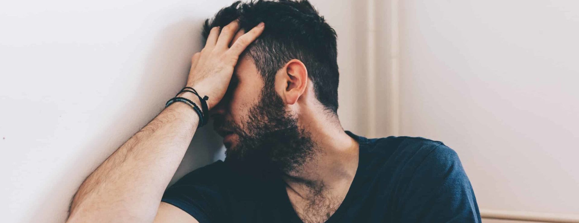 Man with beard and dark hair, suffering from substance use disorder, sitting on floor with head in one hand, covering face