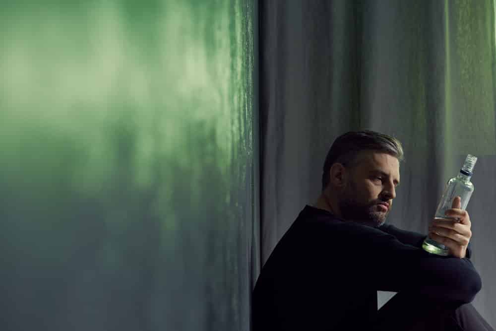 Man in need of alcohol rehab stares at bottle of vodka while sitting on the floor in empty room 