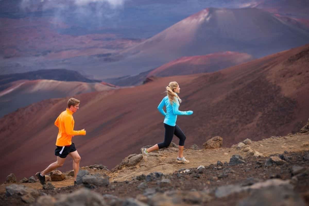 A man and a woman in athletic gear run up a dirt trail with desert mountains as the scenery