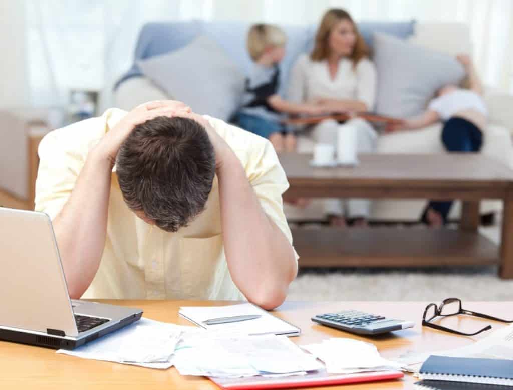 A stressed husband hunched over a table with his laptop and bills; stress can lead to the development of addiction. 