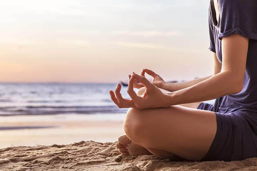 A person sitting on the sand at the beach with their legs crossed in a meditative position. 