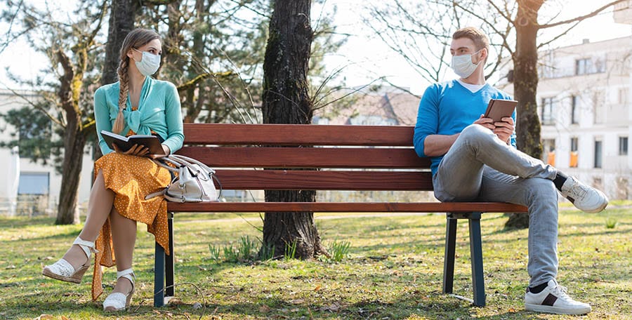 A woman and man, both wearing masks, sit opposite each other on a bench while reading. 