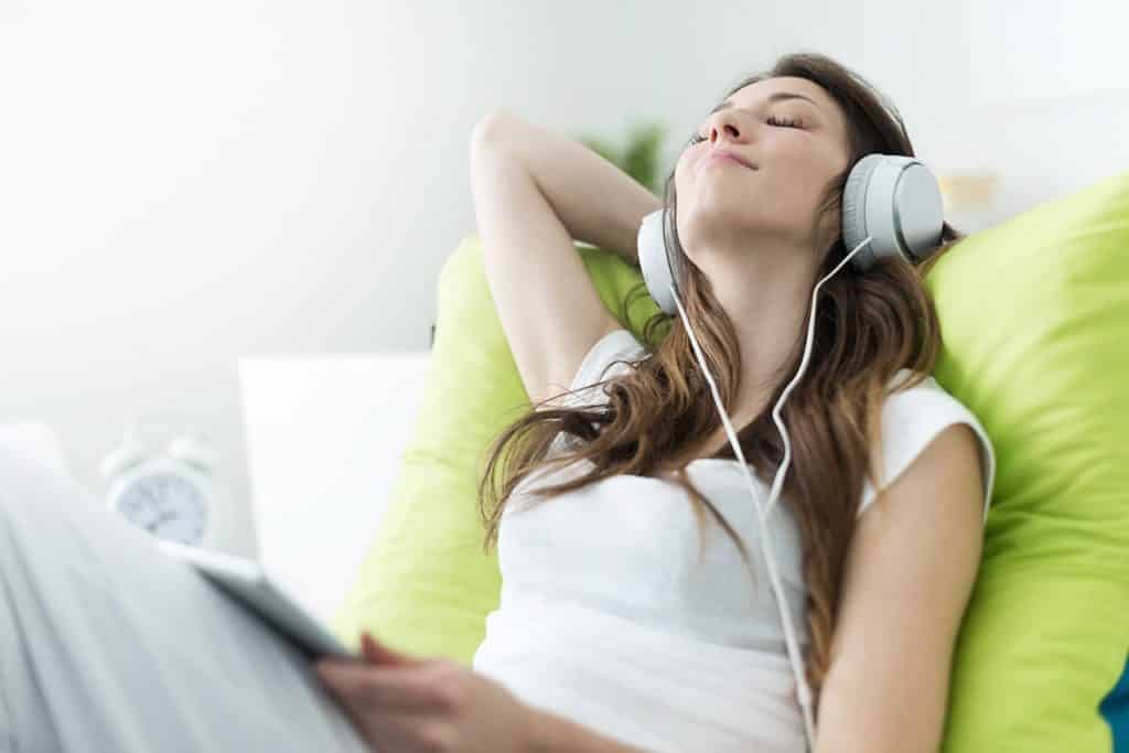 A young woman sitting on her bed with her headphones on is enjoying listening to music. 