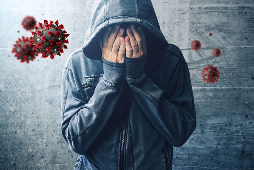 A person in addiction treatment with their hands over their face, surrounded by images of microscopic covid 19 viruses. 