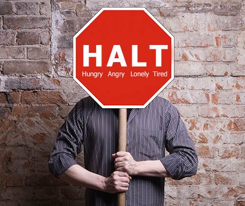 A person is holding up a red sign over his face reading HALT – Hungry, Angry, Lonely, Tired. 