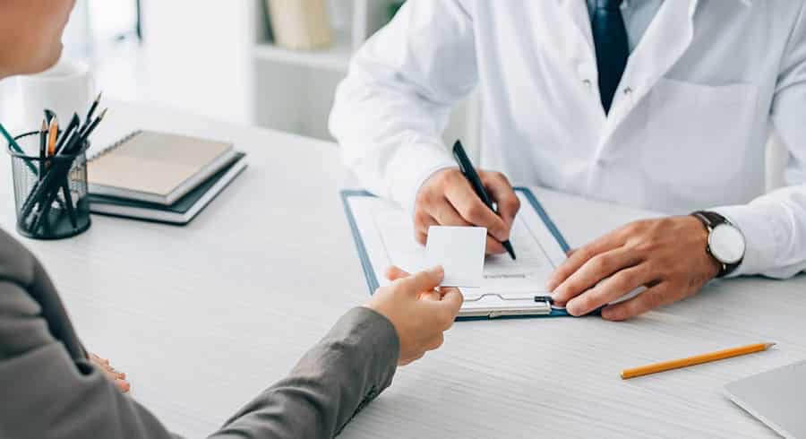 Doctor and patient sit at desk, faces are out of view, doctor is writing on a notepad. Room is brightly lit