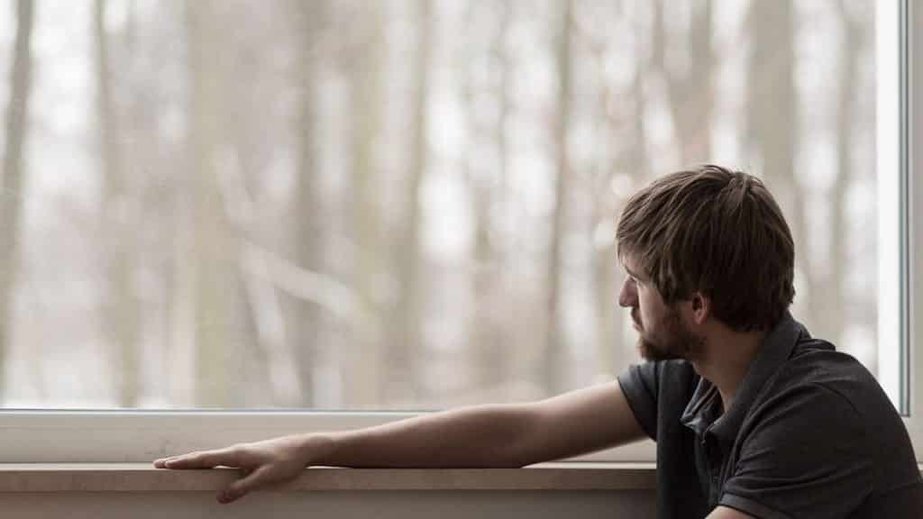 Man feeling the effects of social isolation and its impact on his mental health sits beside a window as he looks out into the yard. 