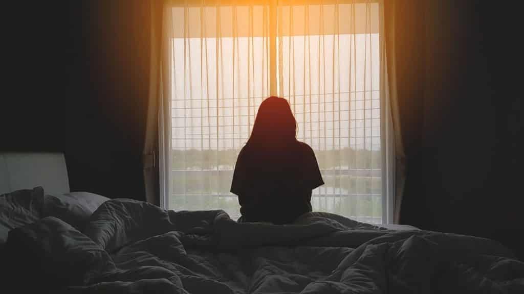 Silhouette of a woman sitting on the end of her bed suffering from social isolation, which is affecting her mental health. 
