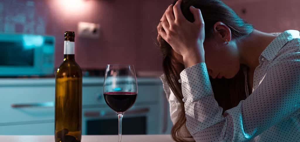 A depressed woman with a open bottle of alcohol and glass of wine in front of her. 