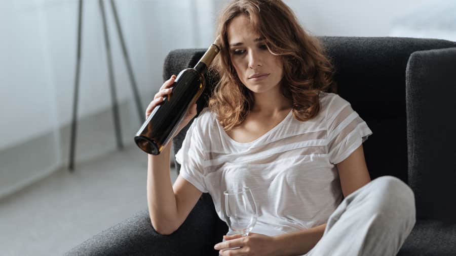 A woman holding a bottle of wine and an empty glass is distraught and reconsidering rehab for her alcohol addiction. 