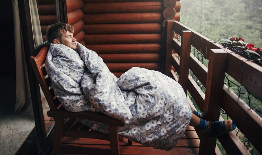 A young content man is sitting on a balcony wrapped up in a blanket looking out to the forest. 