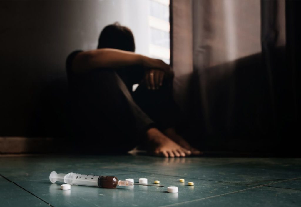 An addict relapsing on drugs during the covid 19 pandemic is sitting on the floor next to various pills and an empty syringe. 