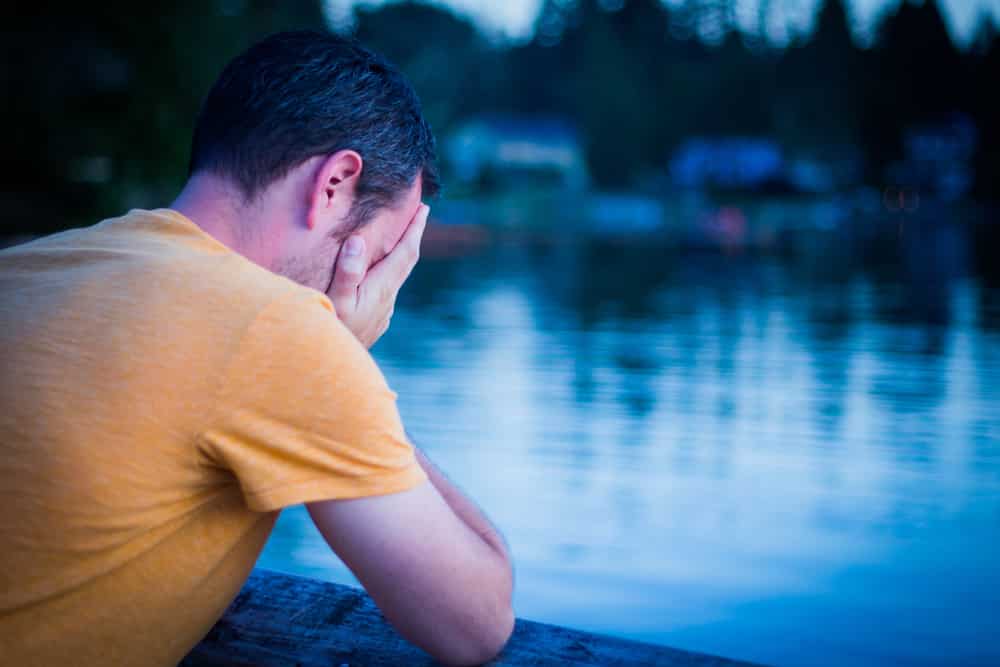 Man in need of addiction treatment in Seattle resting his face in his hands, leaning on fence on bridge overlooking water