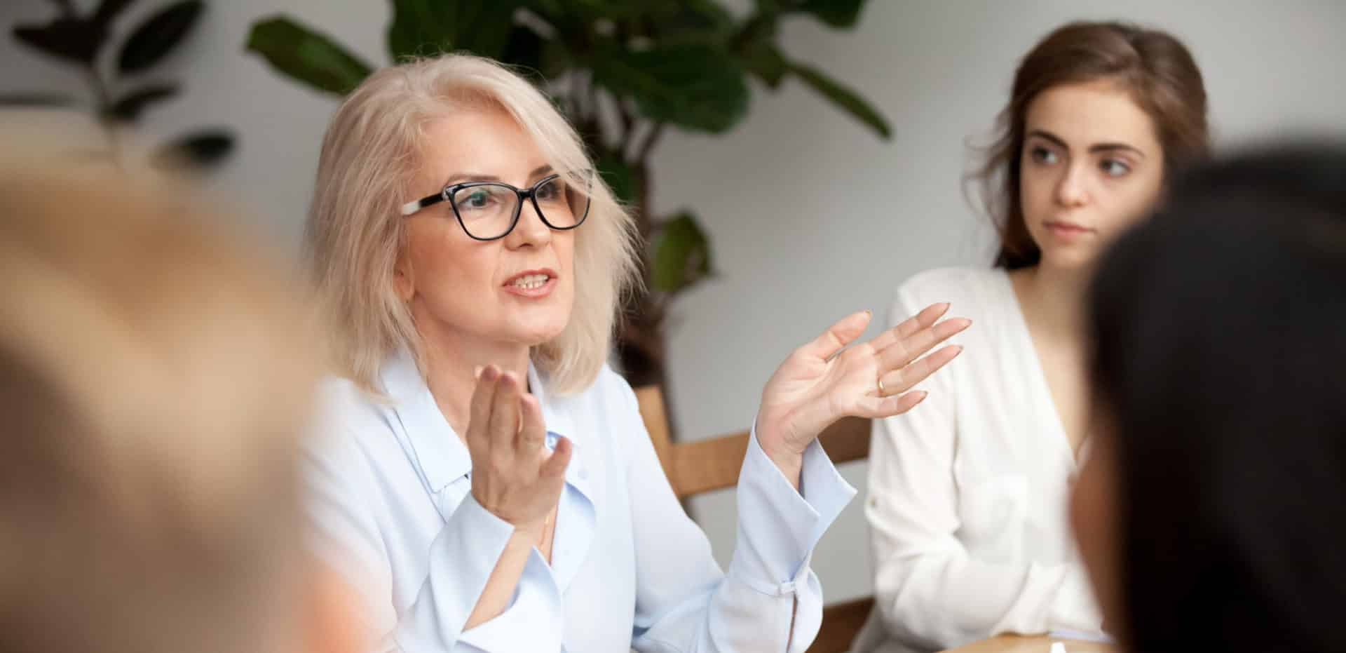 Older woman, professionally dressed with hands out talking to group of adults