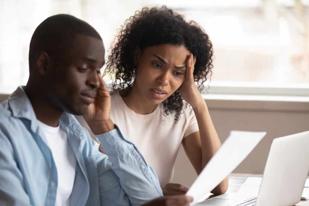 You coupld sitting at home office with computer in front of them with a woman holding a piece of paper, looking confused