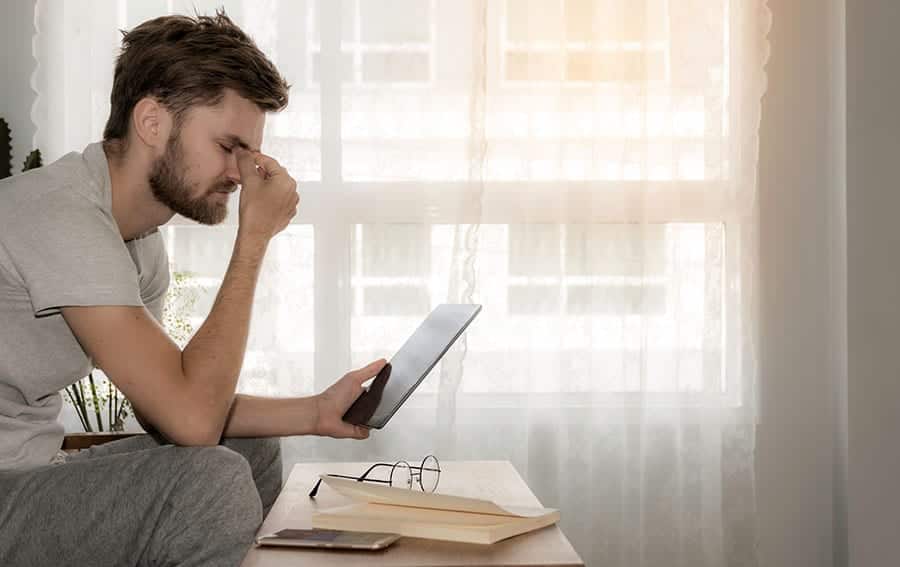 A young man with addiction problems rubs his eyes as he is reading information about addiction rehab on his tablet. 
