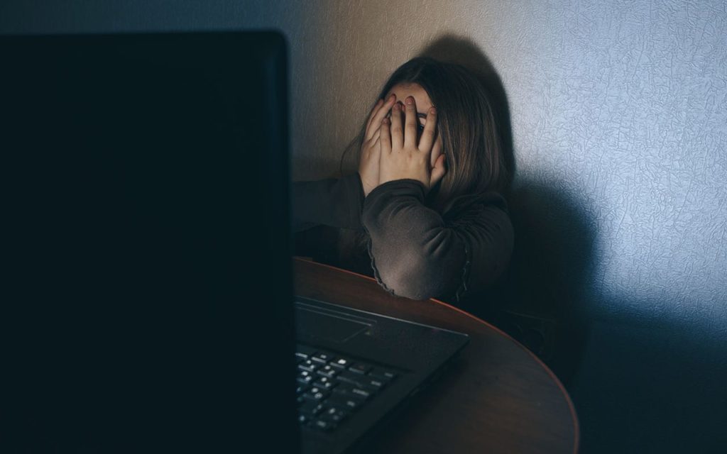 A woman in distress with her hands covering her face while sitting in front of a computer screen contemplates treatment. 