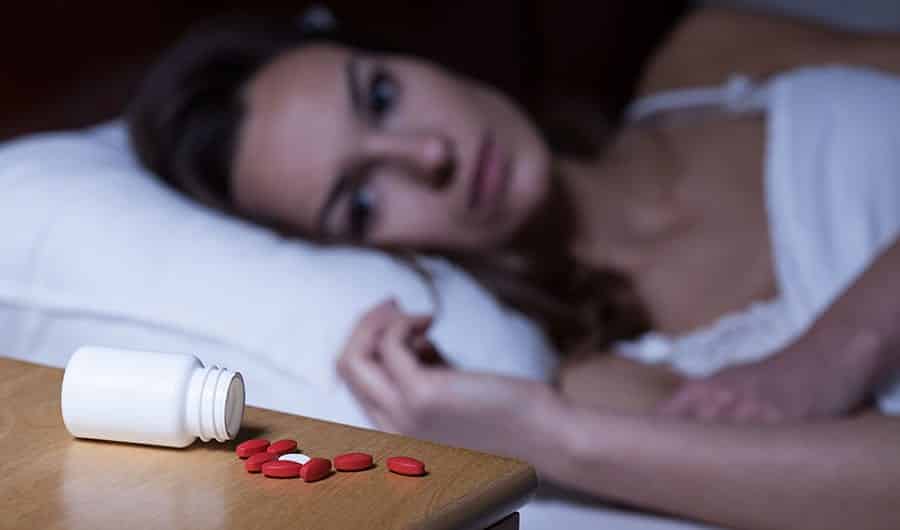 A woman lying in bed is staring at a bottle of prescription pills that have spilled out onto the nightstand. 