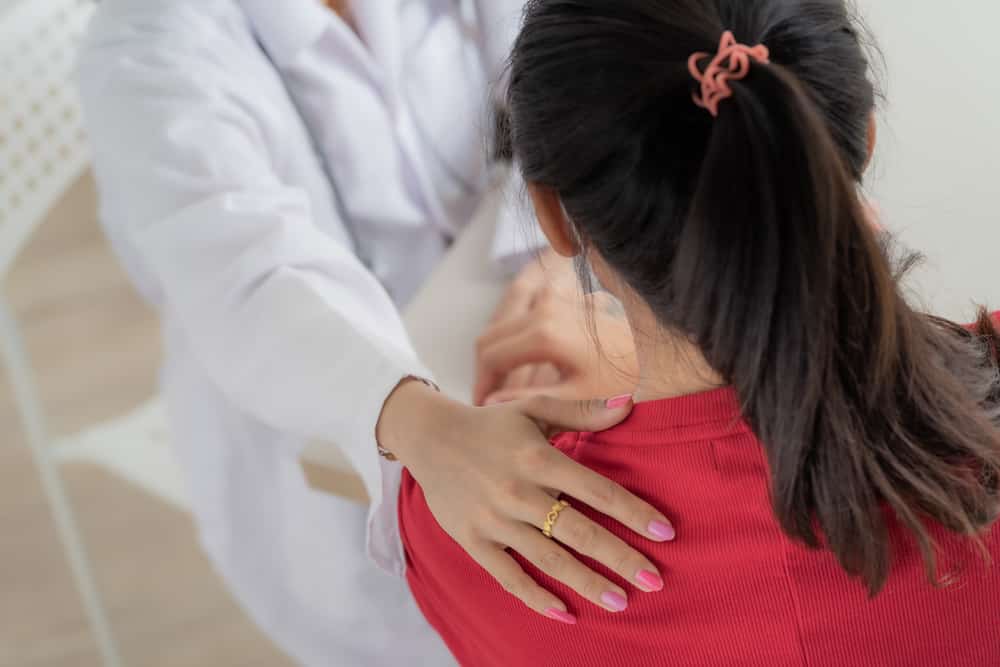 Young girl with head down in red shirt with doctors hand on her shoulder who has pink nails and ring on middle finger