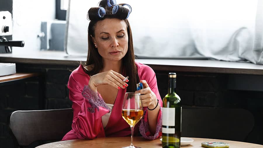 A mother sits in nightgown during the day with a glass of wine and a cigarette - showing she may have a problem with alcohol addiction.