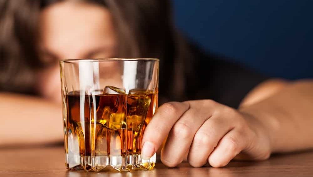 A person is slumped over a table while holding a glass cup of alcohol. 