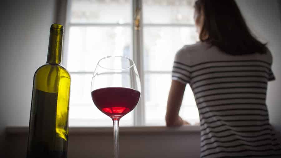A woman suffering from alcoholism looks out a window next to a bottle and glass of wine during the Covid 19 lockdown. 