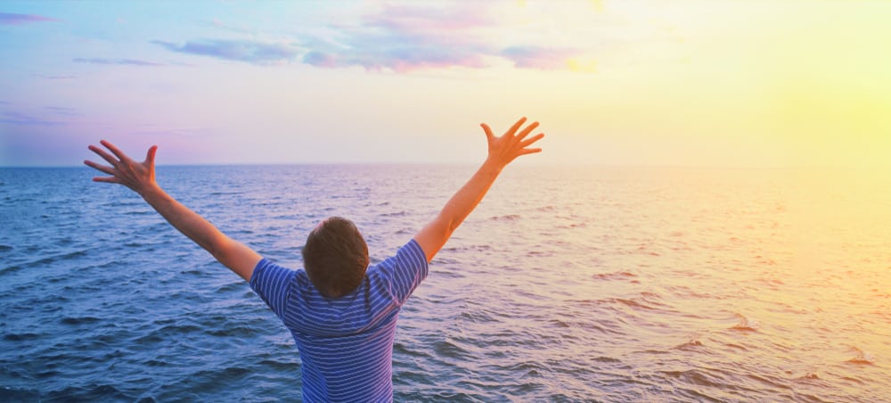 Young mans back, he's facing towards the ocean with arms stretched out and upright, sun is setting- the sky is blue and purple