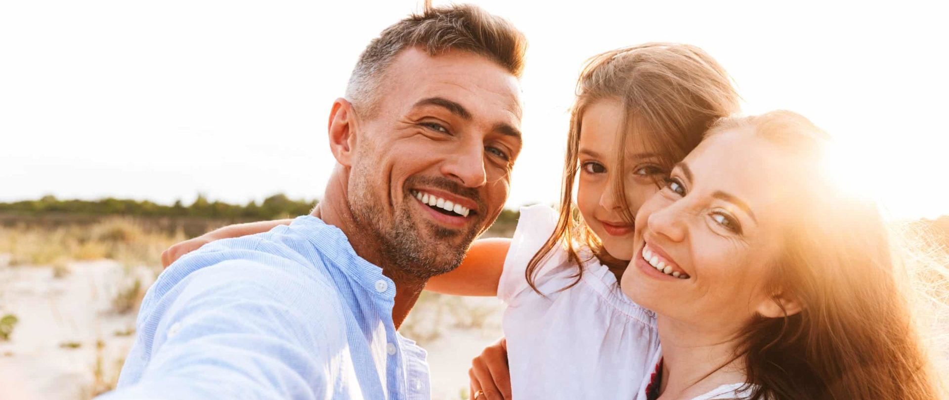 Young parents at the beach with child in the middle of a hug, smiling and staring direct with the sun setting in the background