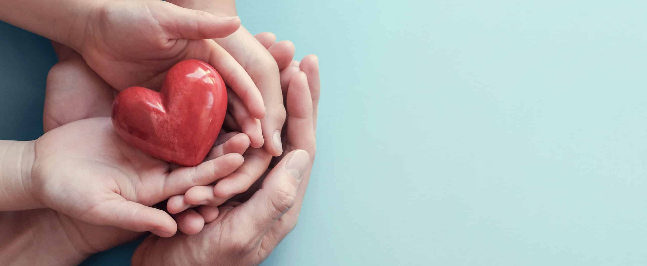 Large hands of man cupped, holding smaller hands of woman cupped, holding child's hands who is holding a ceramic heart