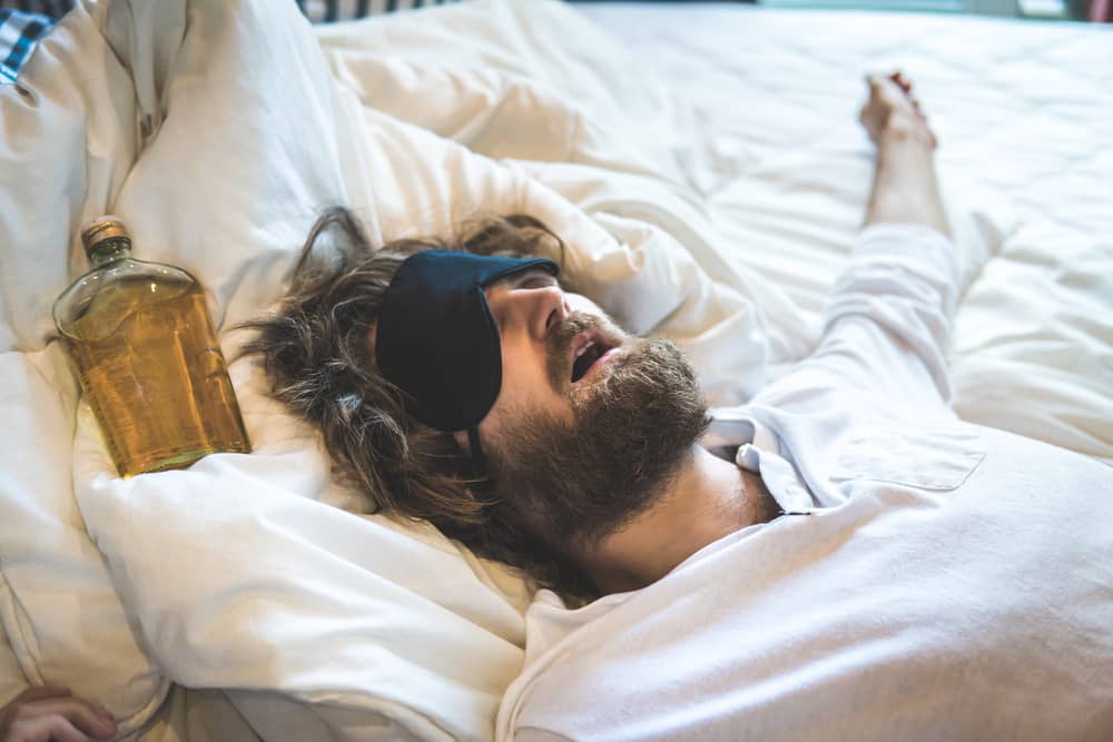 A man lying in bed hungover with a bottle of alcohol next to his head. 
