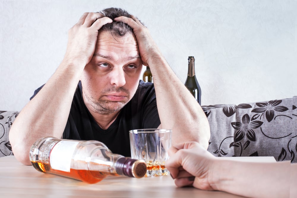 An alcoholic man drunk with his hands on his head sitting at a table with a bottle and glass of alcohol. 