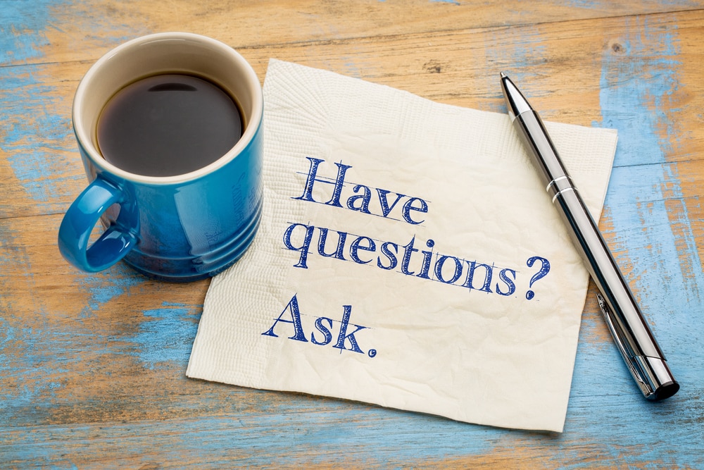 Napkin, pen, and cup of coffee on wood table, the napkin has typed text "Have questions? Ask." on it