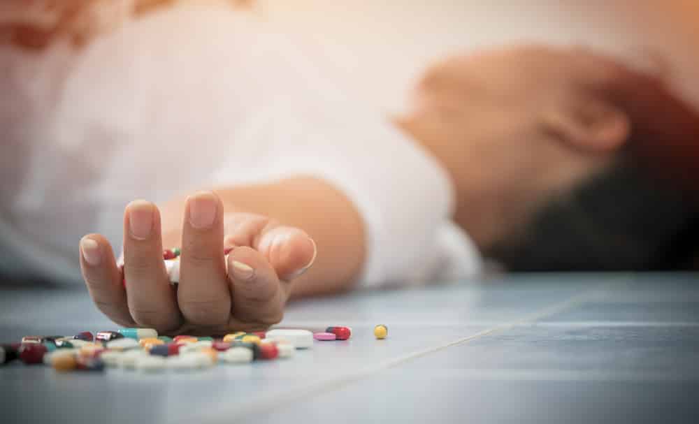 A person who has just overdosed is lying on the floor with various pills spilling out of their hand.