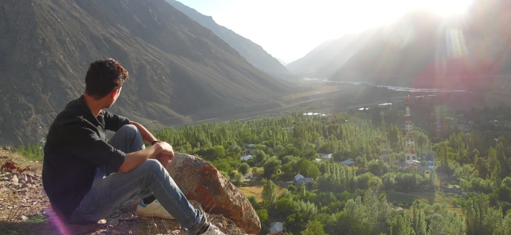 Man sitting down after hiking to a vista point overlooking a valley with mountains surrounding it on a sunny day.