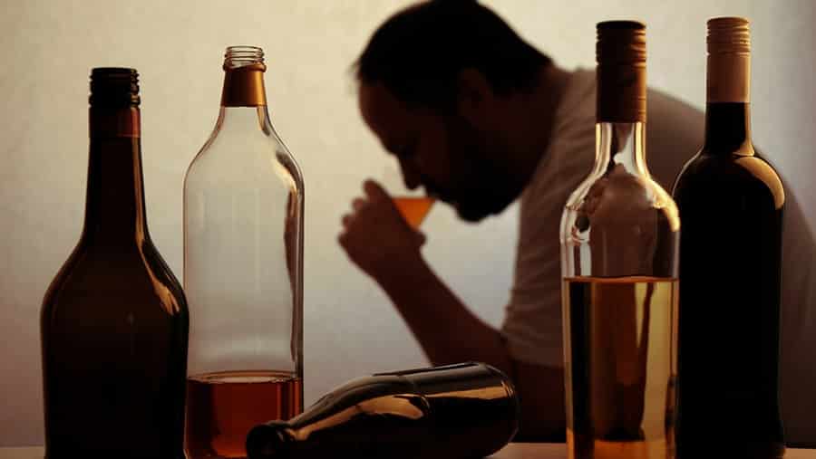 An alcoholic man suffering from alcohol use disorder is drinking a glass of liquor while sitting next to bottles of alcohol. 