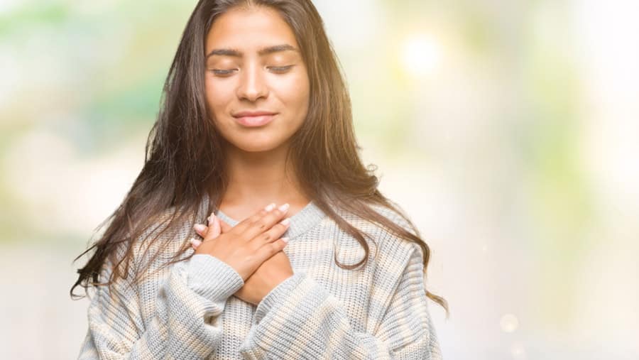 A young woman who is in recovery from addiction has a calm expression with her eyes closed and hands pressed against her chest. 