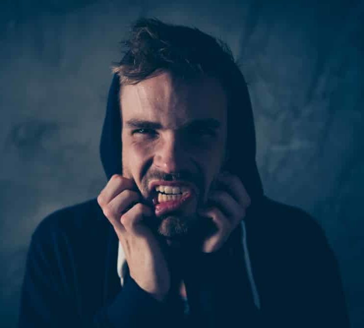 Young Man wearing hoodie in a dark room pulling at his own face in agony