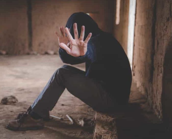 Person sitting in an abandoned dark room, wearing a hoodie with hands up blocking the view of their face