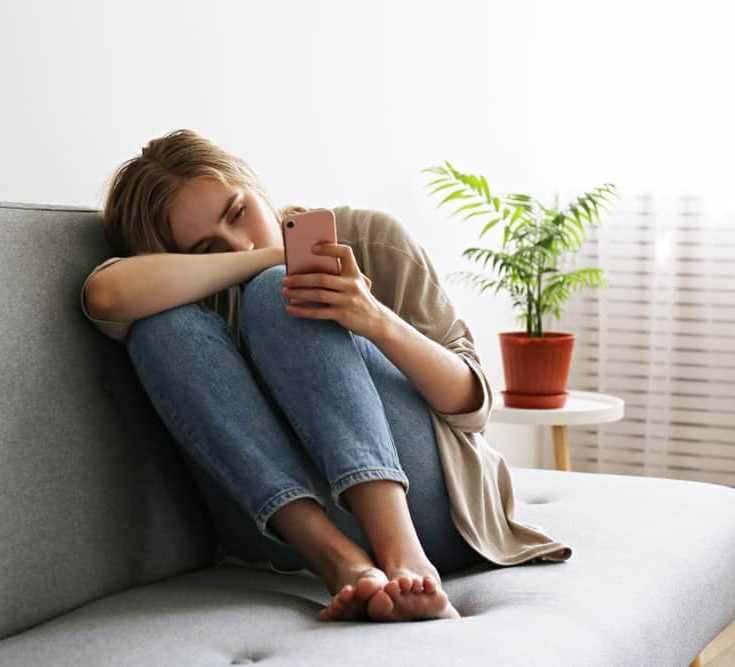 Woman suffering from addiction, hunched over clutching knees staring at phone, on a couch in lightly lit room