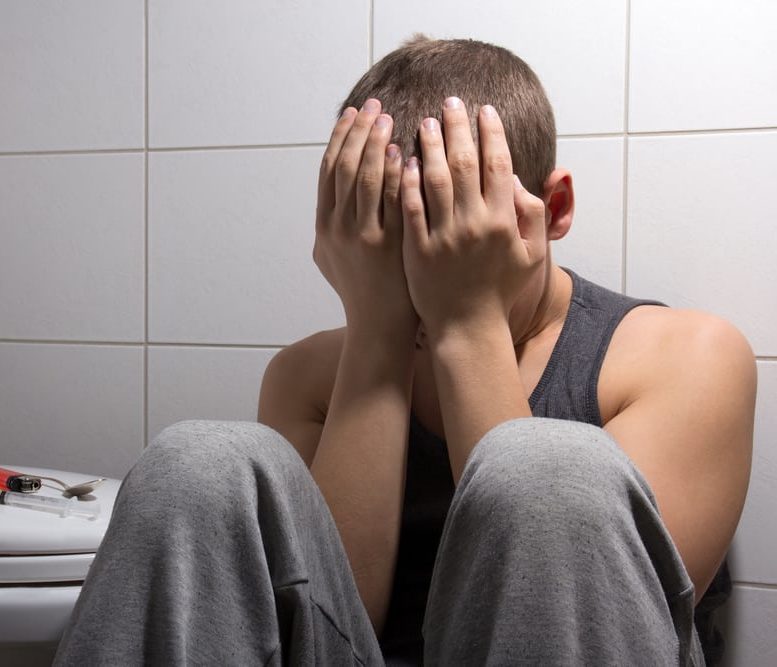 Young man suffering with head in hands, sitting on bathroom floor with needle and spoon sitting on toilet