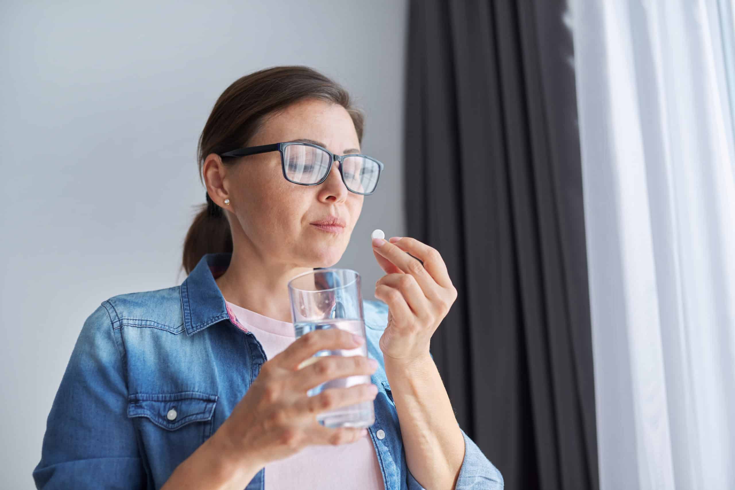 Older woman wearing glasses staring down at ambien, with a glass of water, contemplating if she should take it
