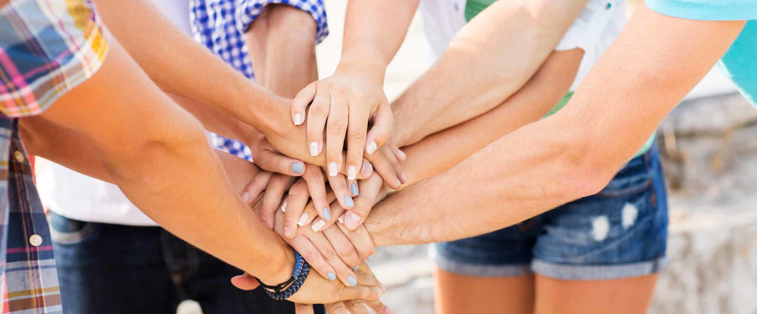 Many hands of men and women on top of each other in group huddle in bright outdoors