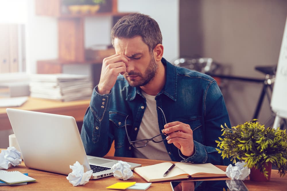 Older man in denim shirt, pinching between his eyes with a migraine sitting at desk