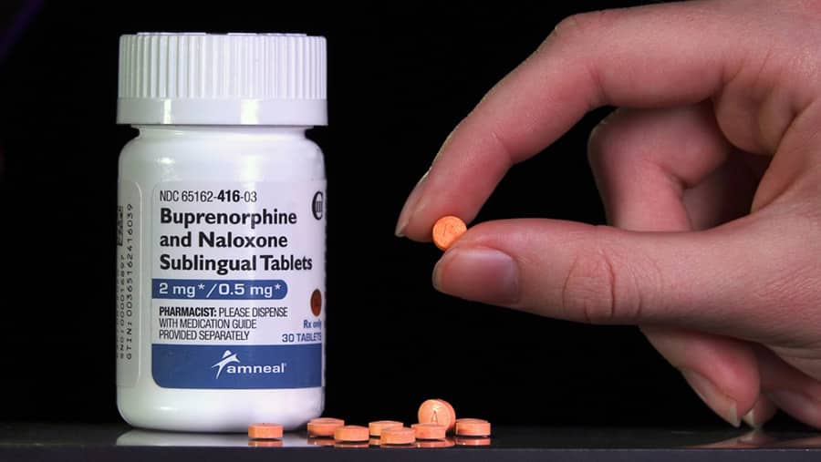 A prescription bottle of suboxone is next to a pile of orange tablets with a hand holding a tablet represents suboxone treatment and withdrawal. 