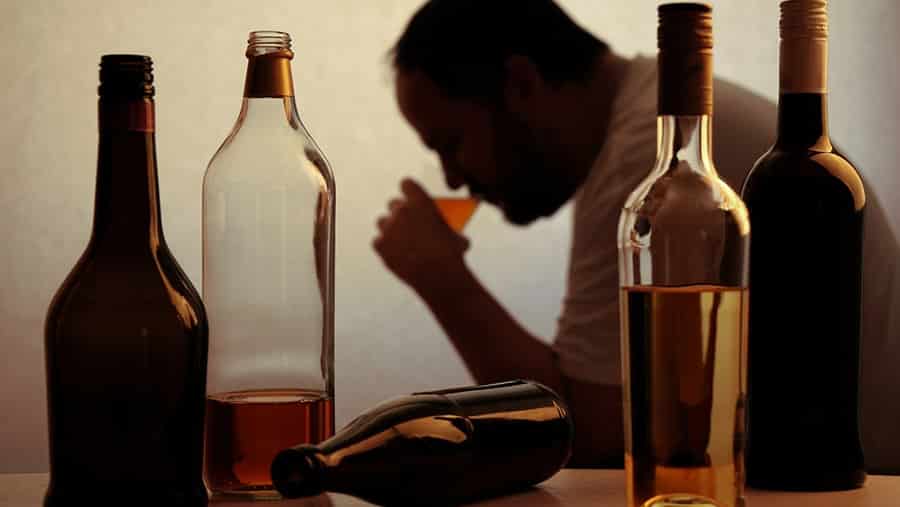 A shadow of a man with early signs of alcoholism and stages of continued abuse is sitting and drinking alcohol in front of various bottles of wine and liquor. 