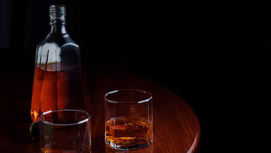 In a dark background, there is a full bottle of alcohol and two glass cups of alcohol on top of a wooden table.